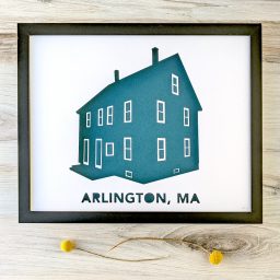 Framed house portrait silhouette depicting a three-story duplex labeled Arlington, MA. Silhouette has a teal background, and frame is sitting on a woodgrain background.