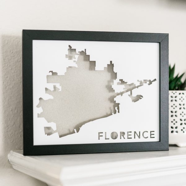 Custom framed paper art map of Florence, AL with a light gray background on a white mantle near a small potted plant