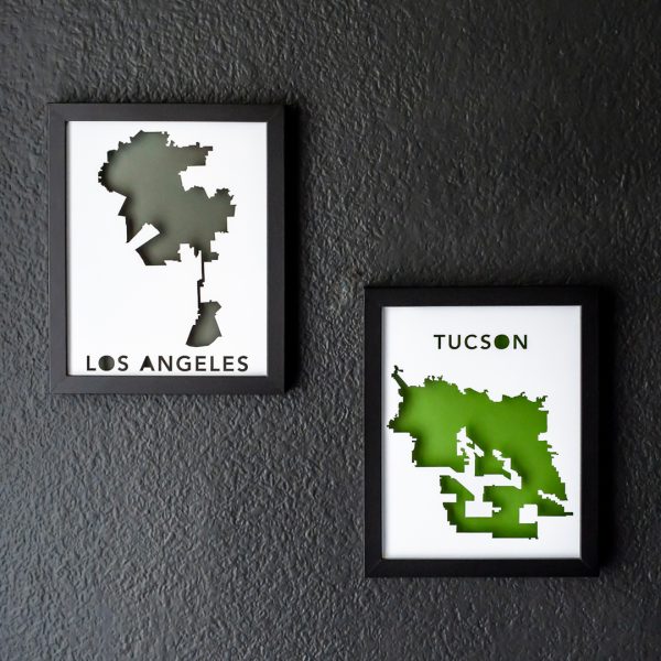 Framed rectangular map of Los Angeles, CA with a gray background and Tucson, AZ with a green background. Black frames hang on a dark gray wall with dramatic lighting from the side.
